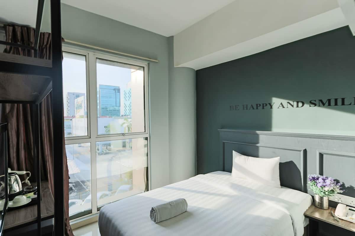 Una habitación de hotel con un ventanal grande, una cama con ropa blanca, una pared oscura con una frase motivacional y una mesita con un jarrón de flores.