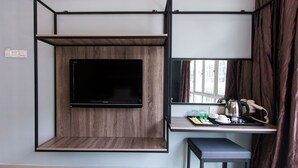 Ein kompaktes Zimmer mit einem an der Wand befestigten Flachbildfernseher, einem kleinen Tisch mit Wasserkocher, Tassen und Gläsern sowie einer Sitzbank.