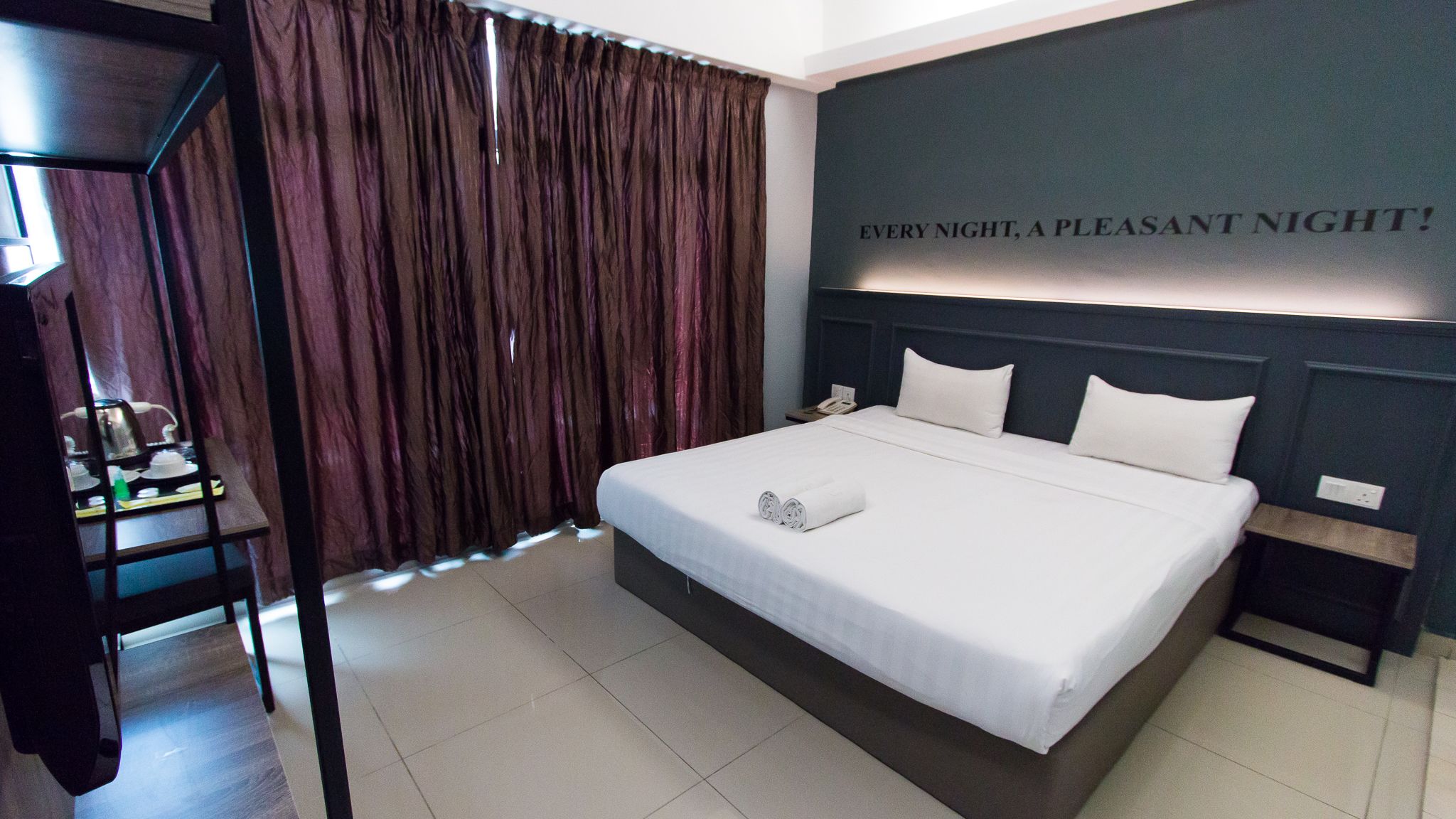 Una cama bien hecha con un almohadón y una toalla enrollada, una mesita de noche con un teléfono, y una pared con un letrero que dice "EVERY NIGHT, A PLEASANT NIGHT!".
