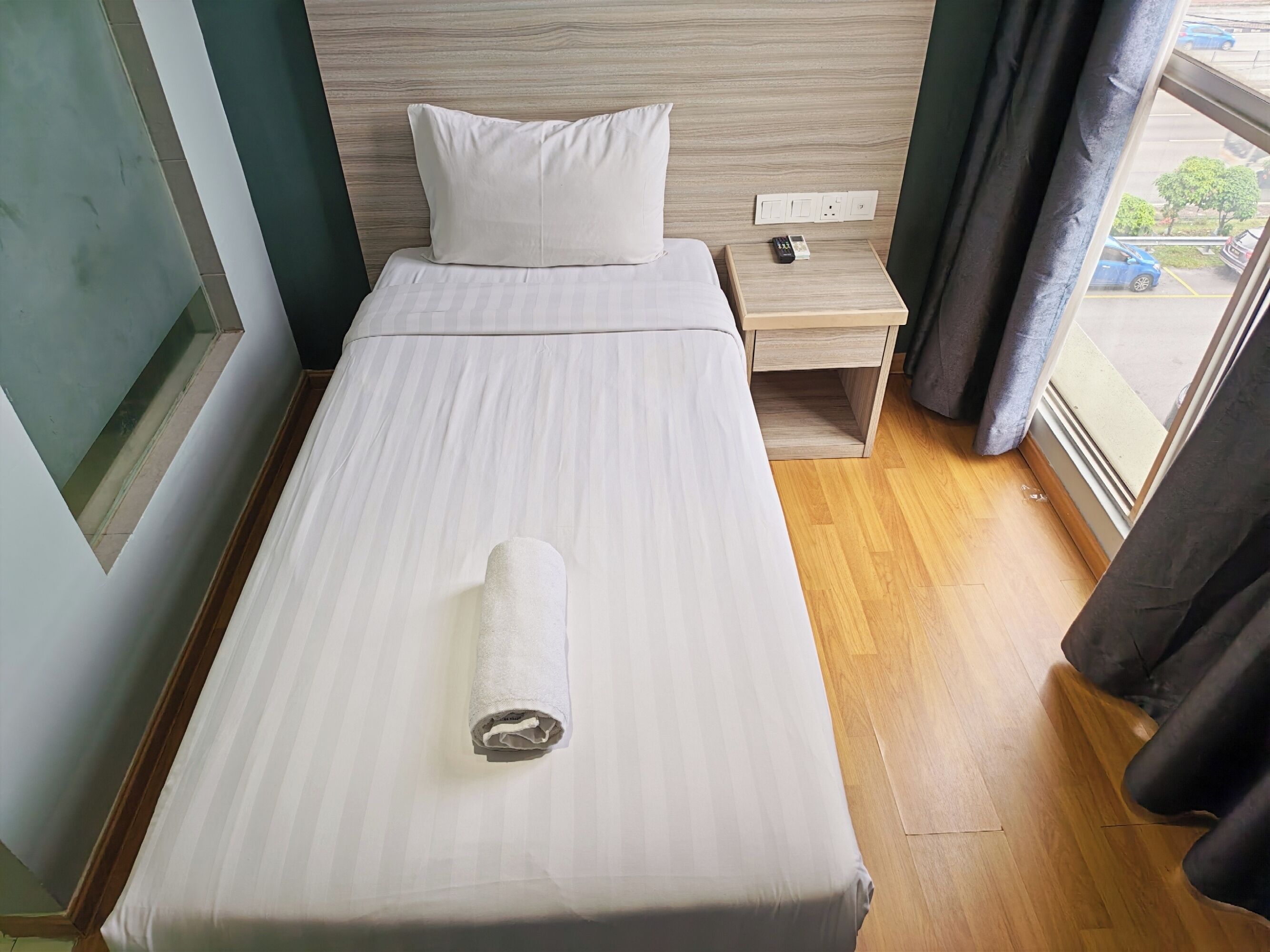 A neatly made bed with a rolled towel, a bedside table with a phone and a power outlet, and a window with a view of the street.