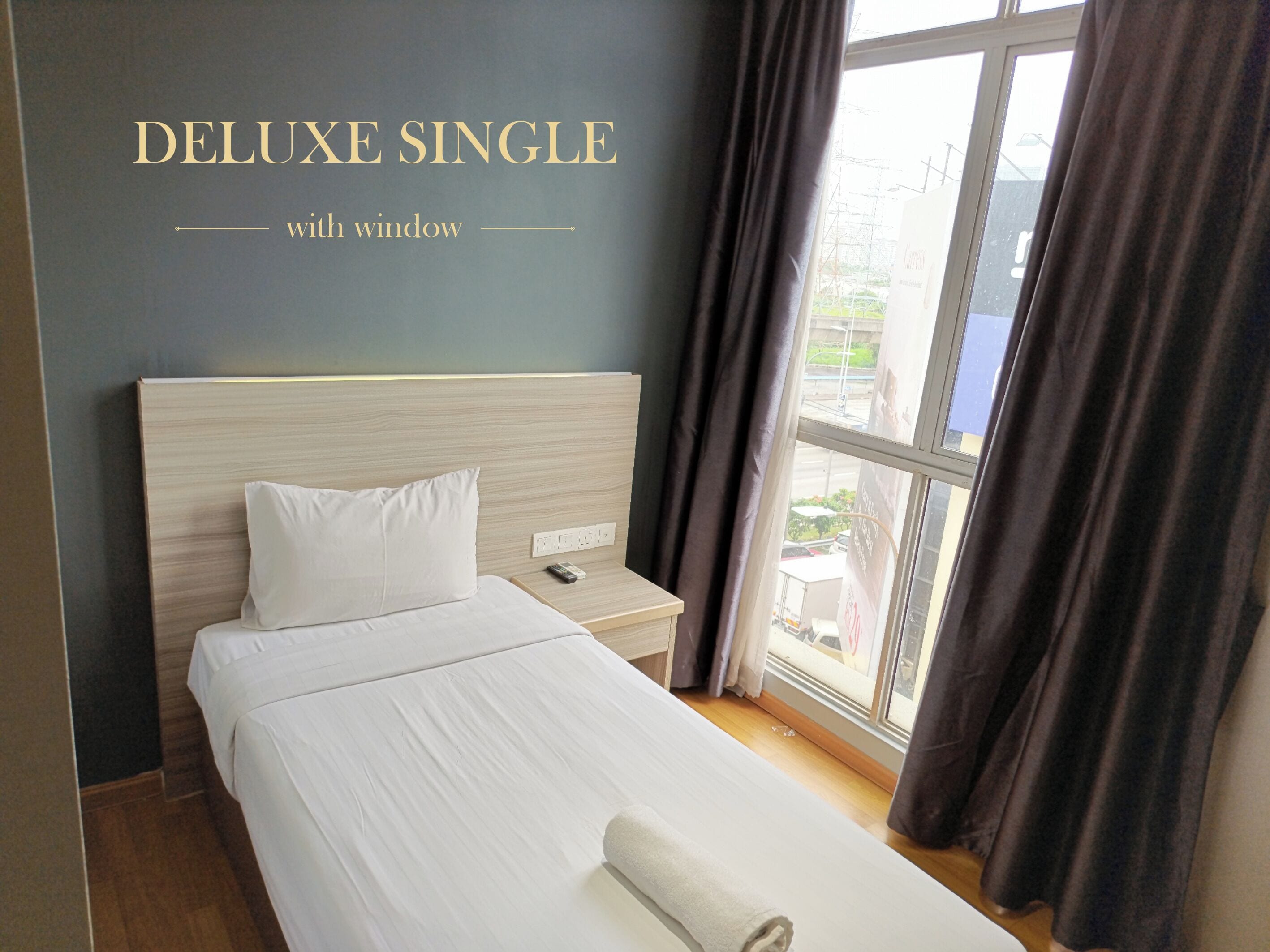 A DELUXE SINGLE room with a window, a wooden headboard, a white bed, a bedside table with a remote control, and a rolled white towel.