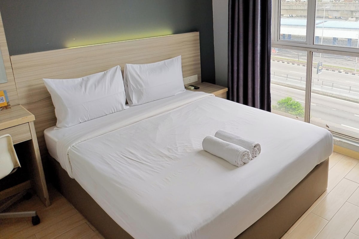 A hotel room with a large bed, a bedside table, a chair, and a window with a view of the street.
