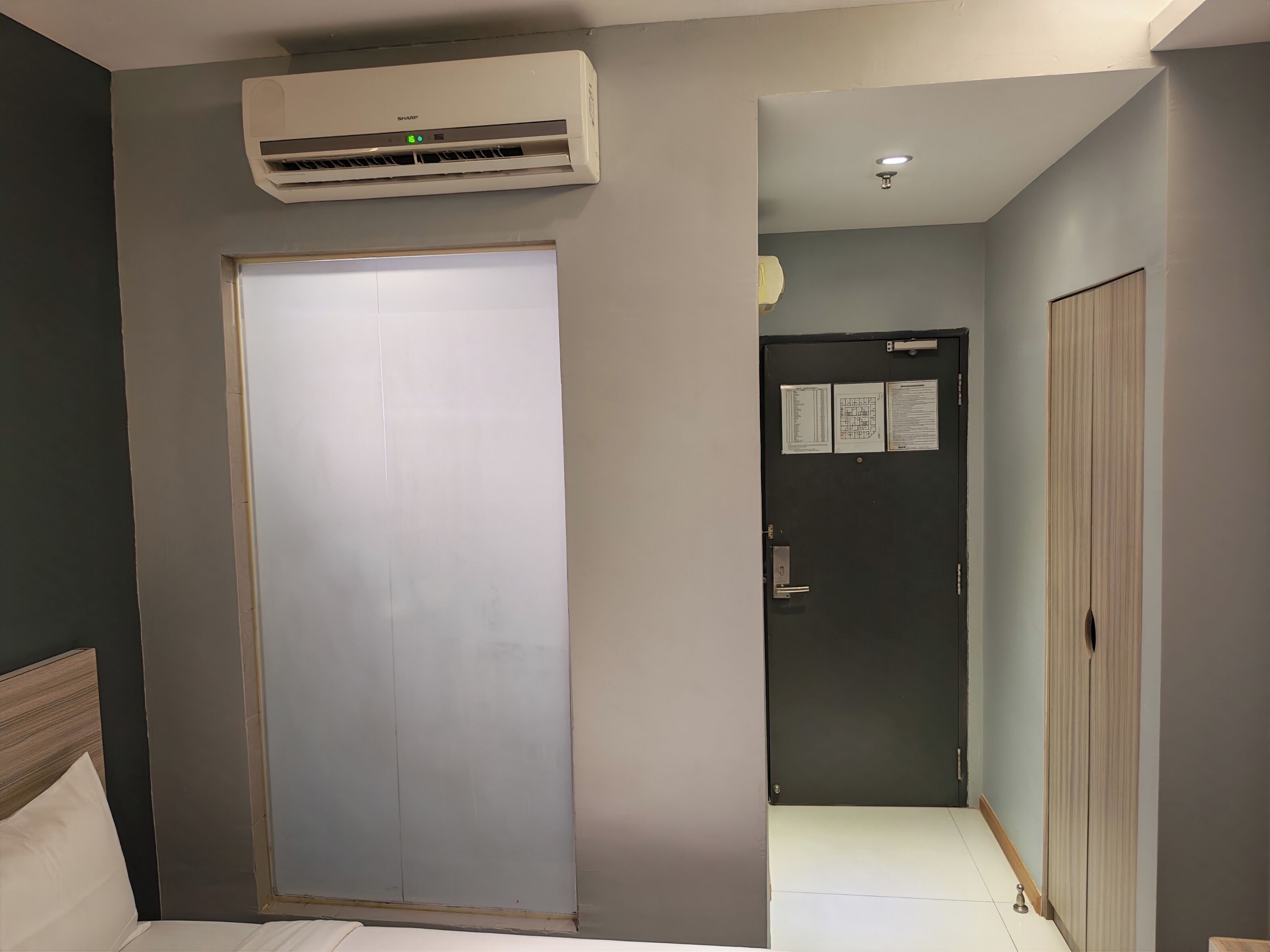 A hotel room with a large wardrobe, a bed with white bedding, a wall-mounted air conditioner, and a door with a notice.
