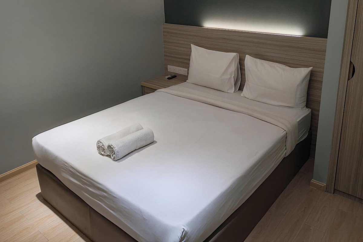 A neatly made bed with a white duvet and a rolled towel, a wooden headboard, and a nightstand with a phone.