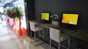 A reception area with two computer monitors, a keyboard, and a mouse on a desk. There are two white chairs in front of the desk. A large potted plant with red flowers is on the left side of the desk.