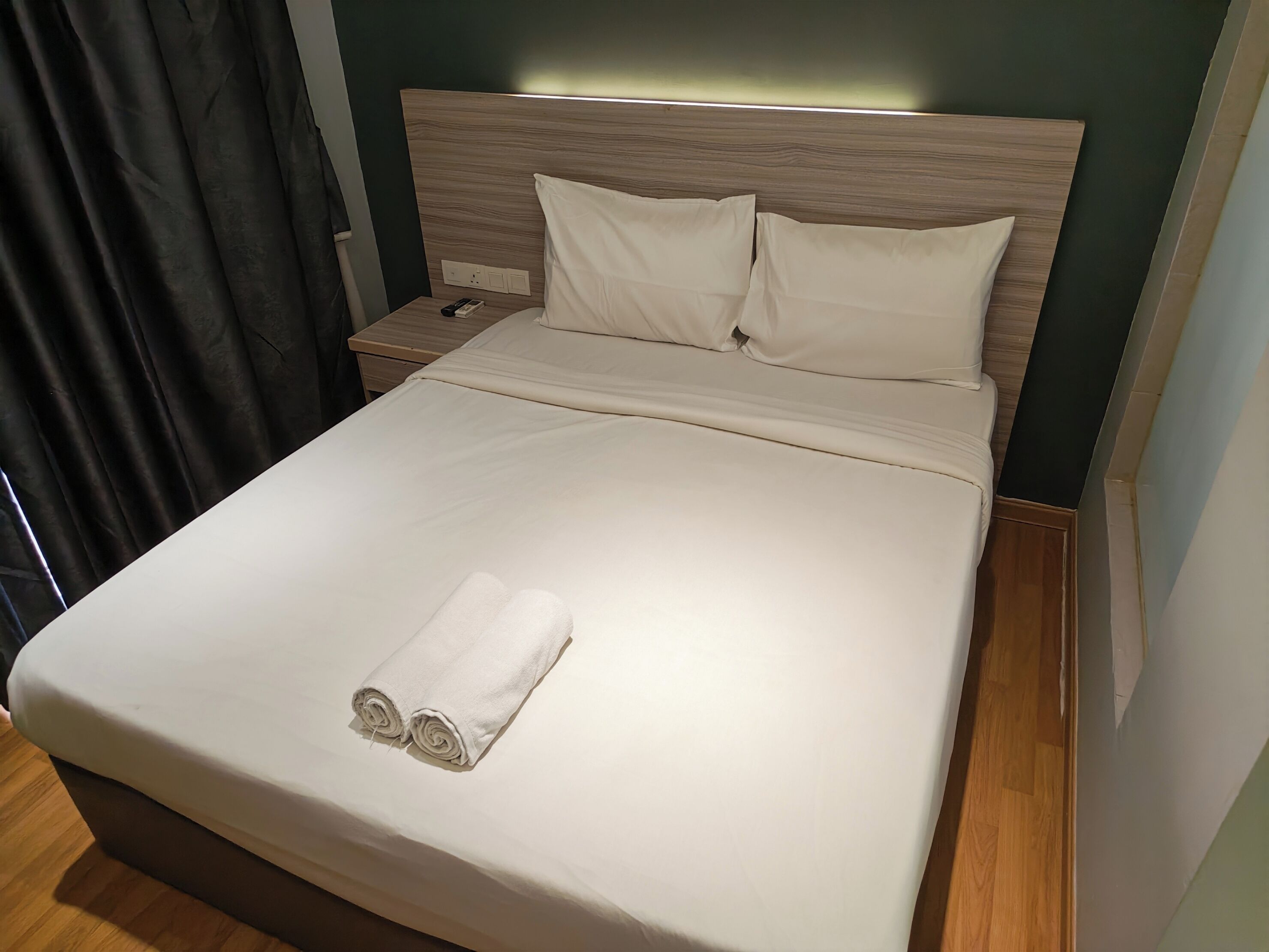 A neatly made bed with a white duvet, a wooden headboard, and a remote control on the nightstand beside it.