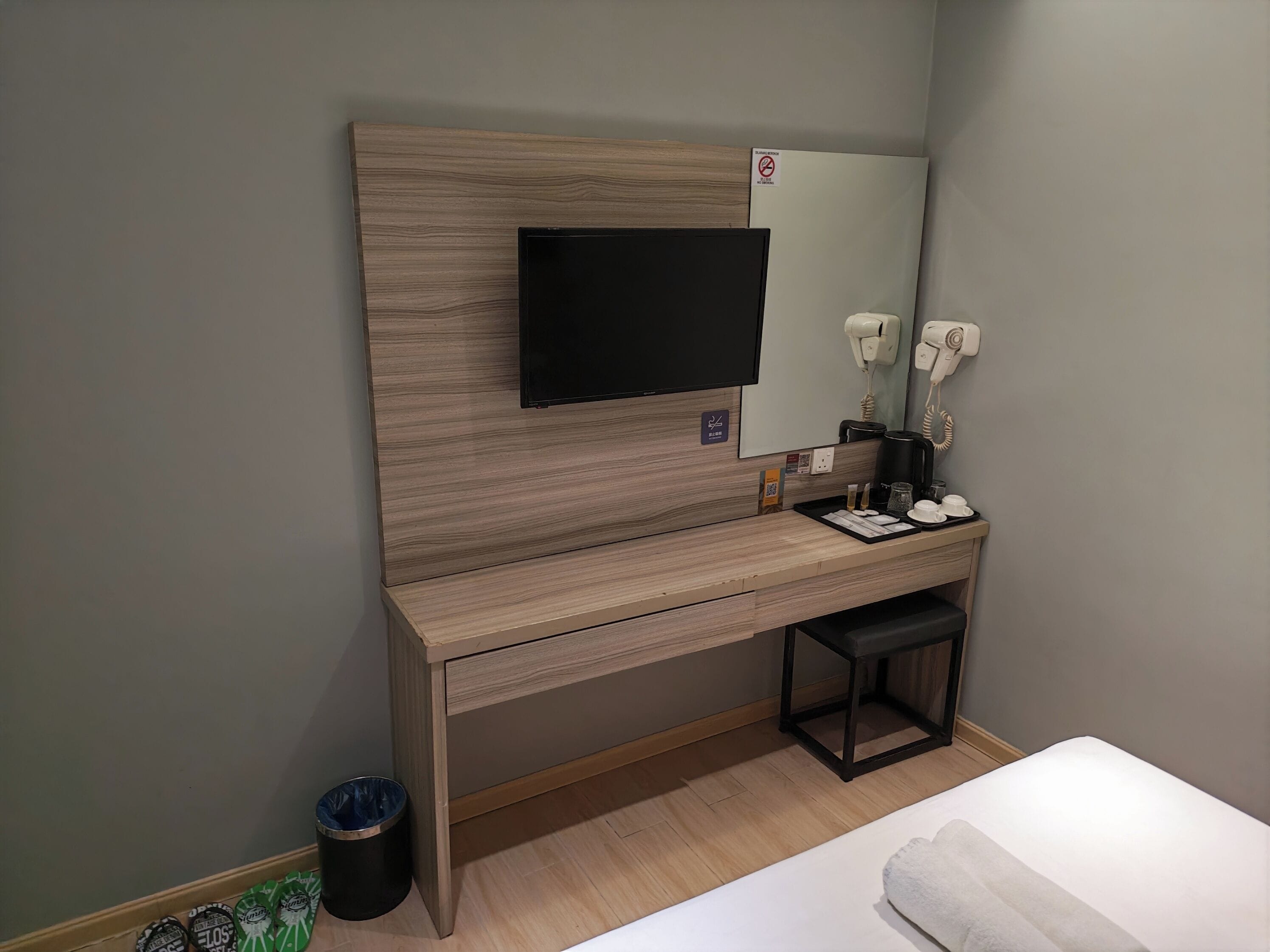 A hotel room with a wooden desk, a flat-screen TV, a mirror, a hairdryer, a coffee machine, and a trash bin.