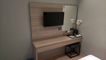 A hotel room with a wooden desk, a flat-screen TV, a mirror, a hairdryer, a coffee machine, and a trash bin.