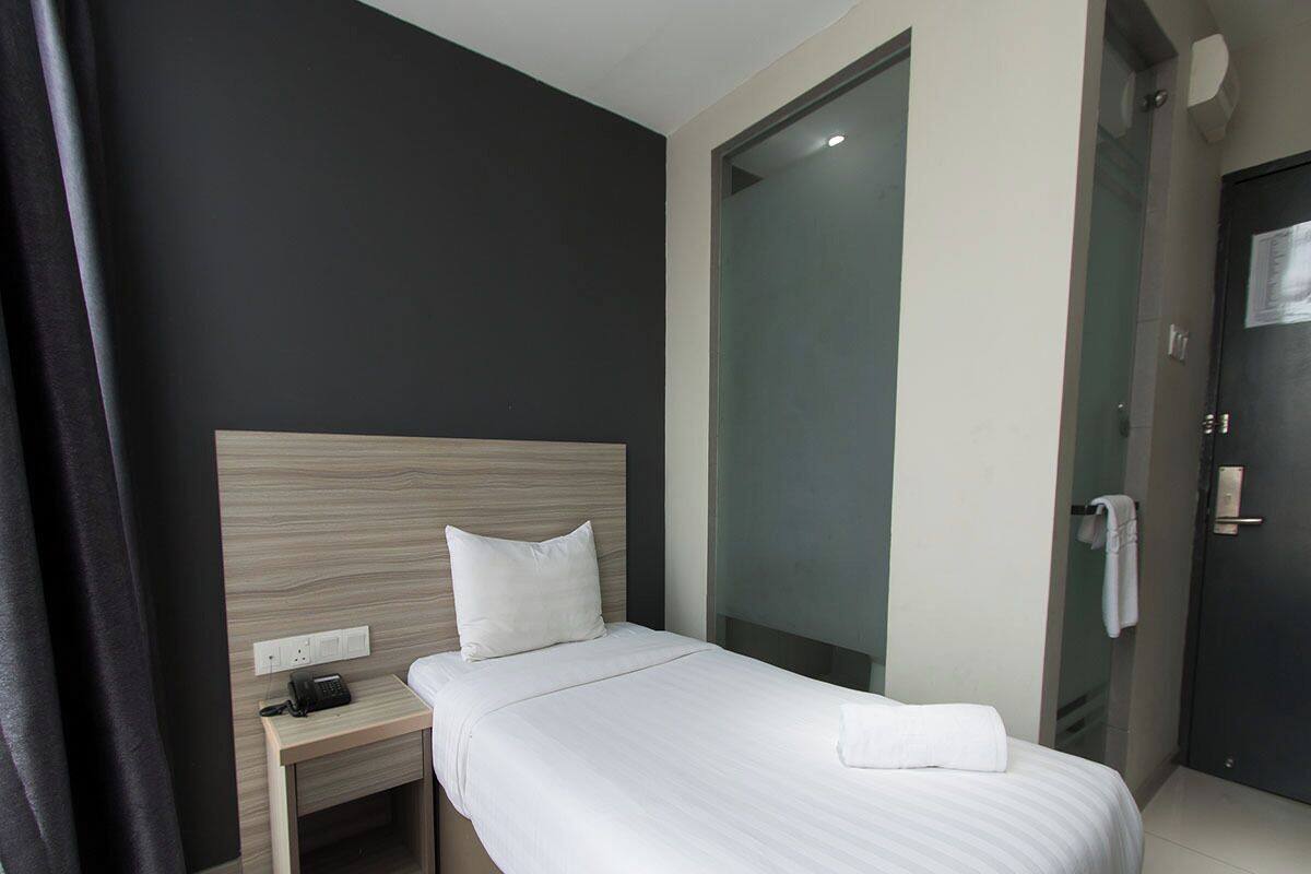 Habitación de hotel con cama, mesita de noche con teléfono, cabina de ducha de vidrio y una puerta con una toalla colgada.