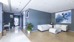 A modern reception area with a white reception desk, a seating area with white sofas, a large blue floral painting on the wall, and a potted plant.