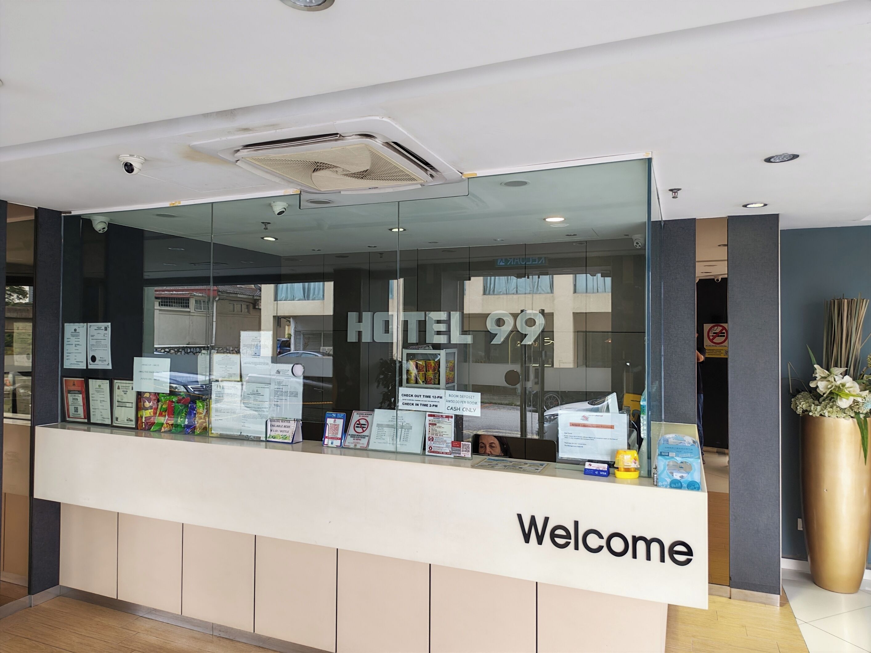 A hotel reception desk with a 'Welcome' sign, a glass partition, and various informational brochures.
