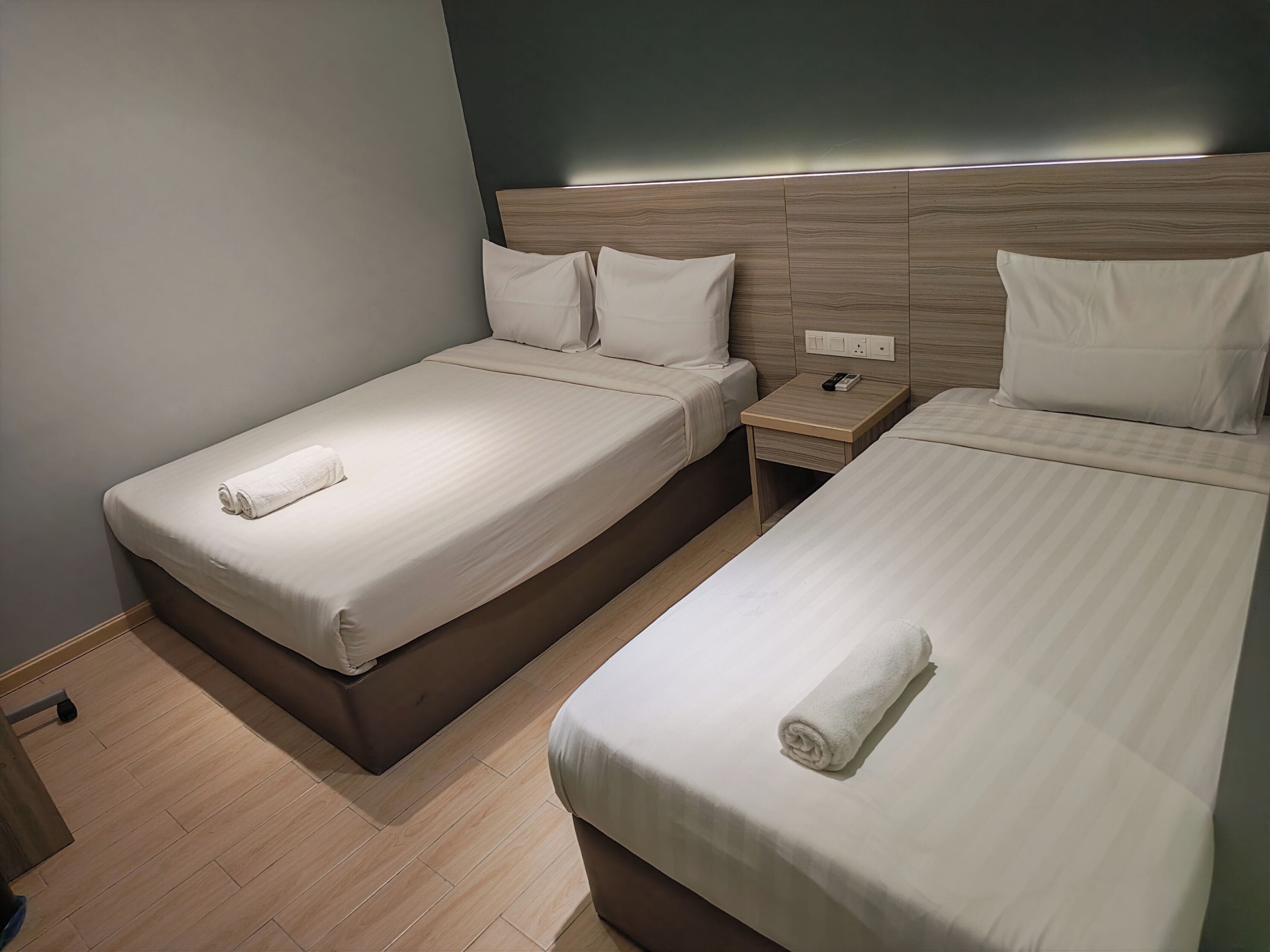 A hotel room with two single beds, each with a neatly folded towel, a wooden headboard, and a bedside table with a phone.