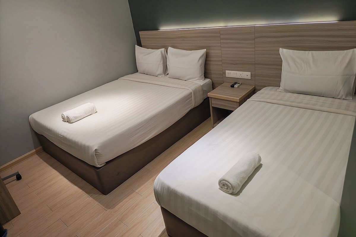 A hotel room with two single beds, each with a neatly folded towel, a wooden headboard, and a bedside table with a phone.
