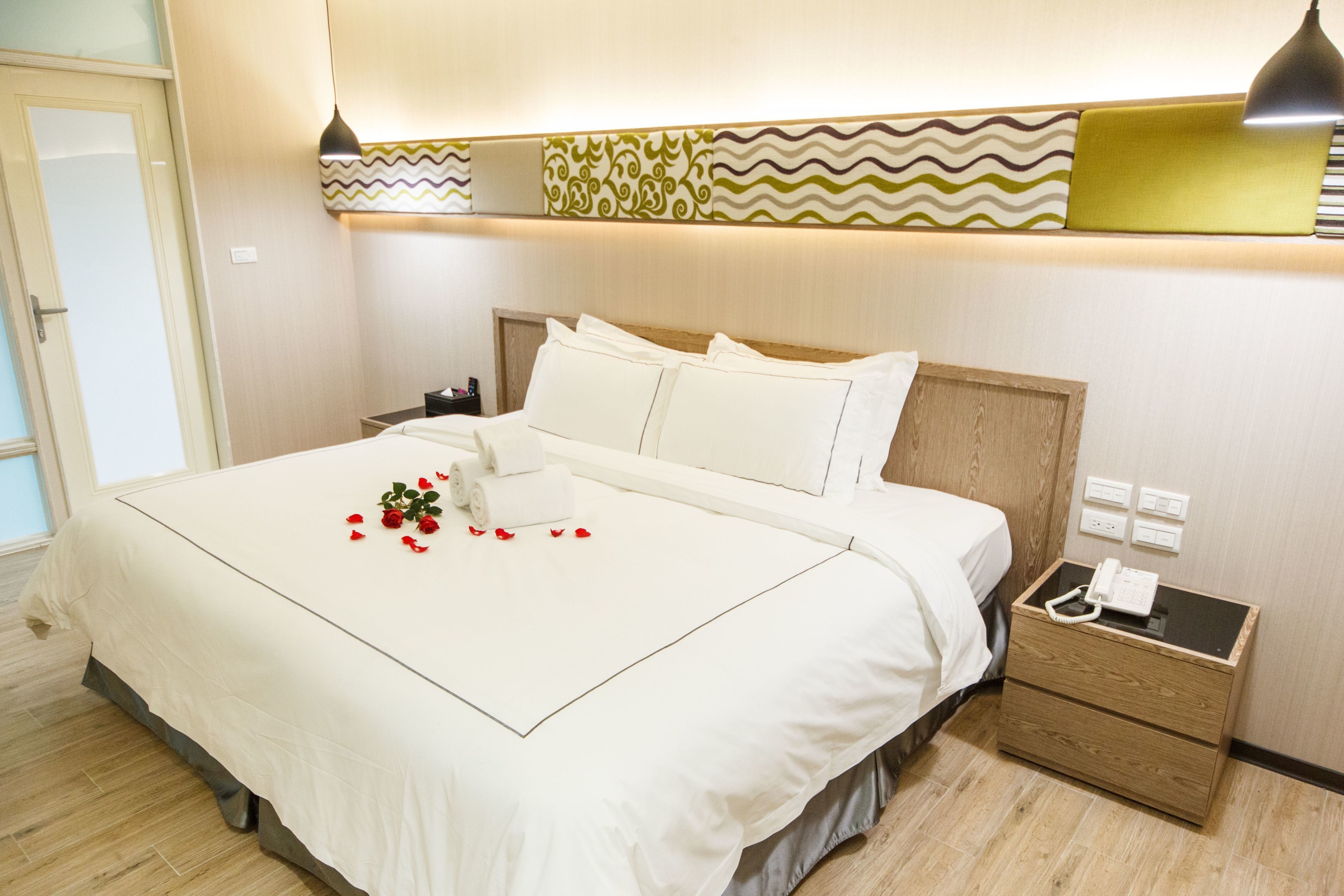 A neatly made bed with white linens and a decorative pillow arrangement. A bedside table with a lamp and a phone is visible.