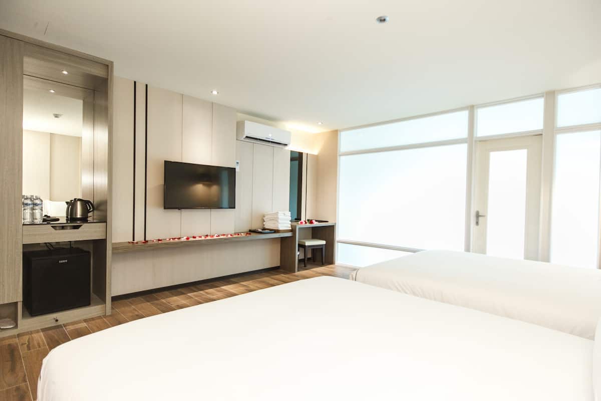 A hotel room with a large bed, a TV mounted on the wall, a small desk with a kettle, and a window providing a view of the outside.