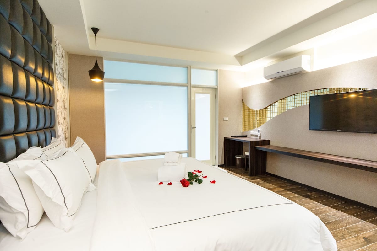 A modern hotel room with a large bed, a flat-screen TV mounted on the wall, and a view of the city through the window.
