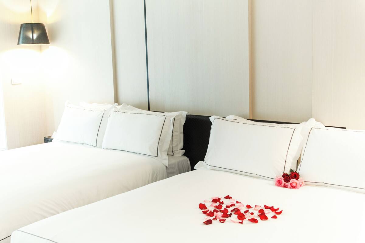A hotel room with two beds, white bedding, a wall-mounted lamp, and rose petals on the bed.