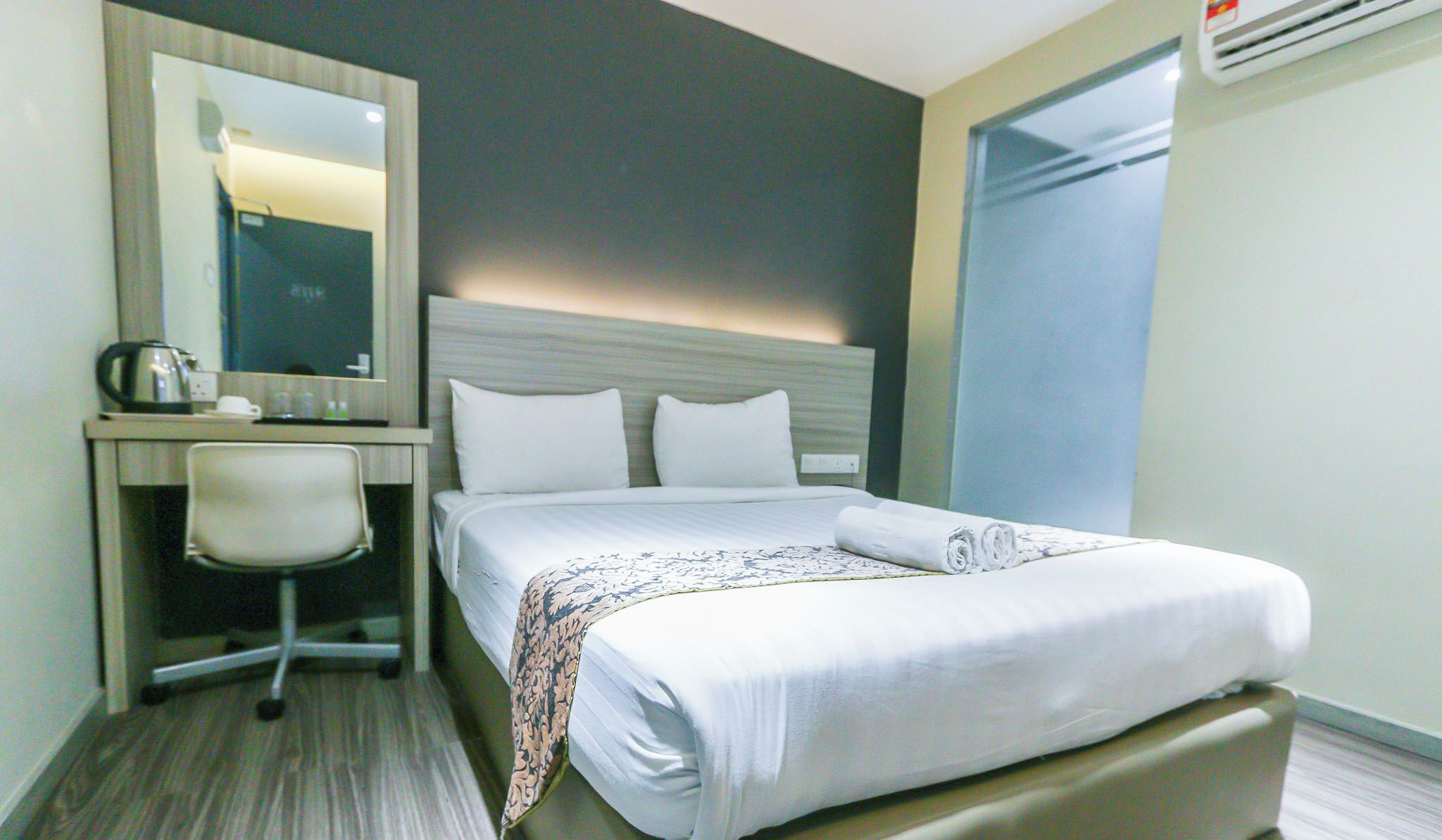 A hotel room with a bed, a desk with a chair, a mirror, and an air conditioning unit.