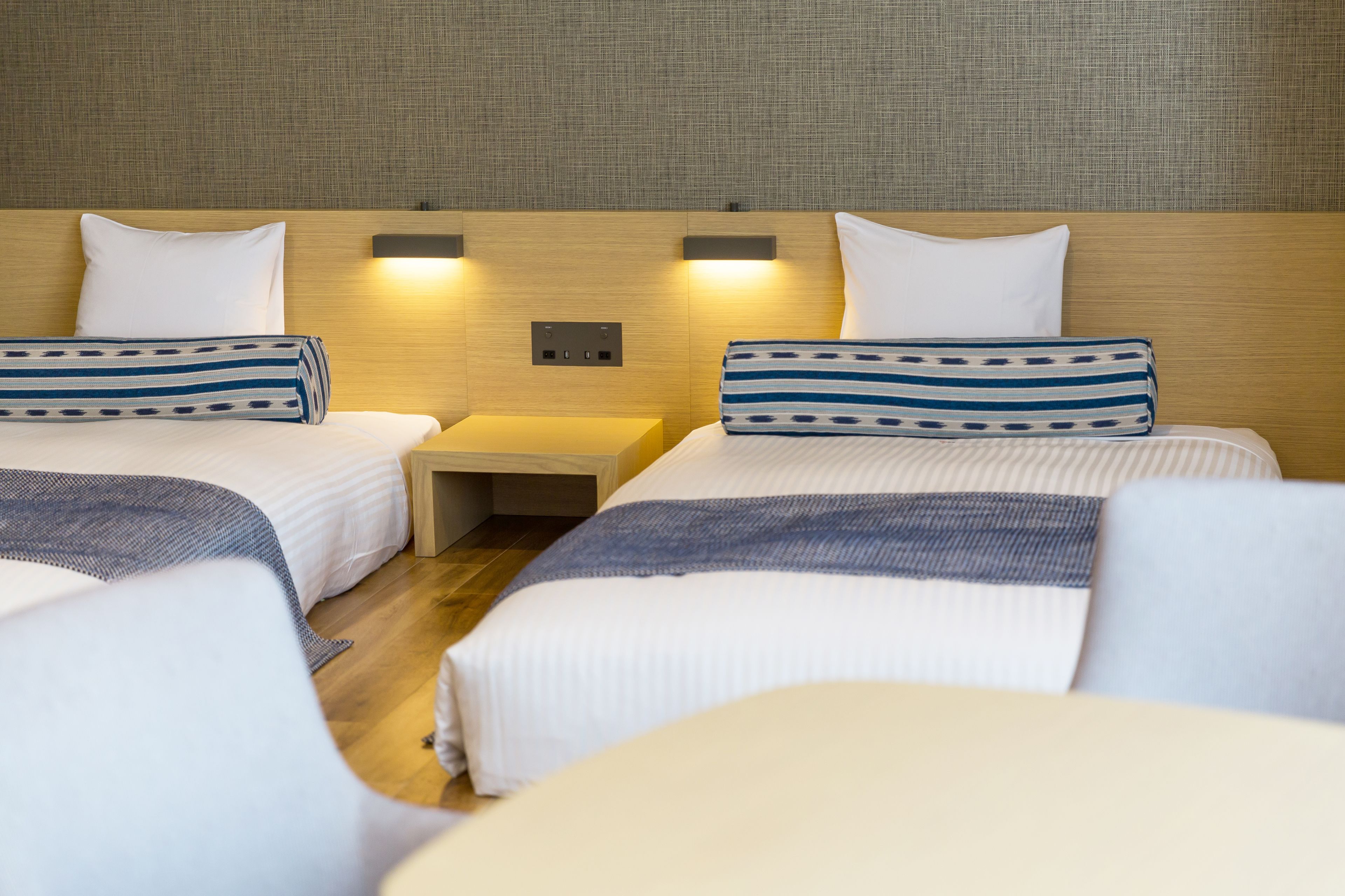 A hotel room with two beds, each with a pillow and a striped blanket. There is a nightstand between the beds with a lamp on it.