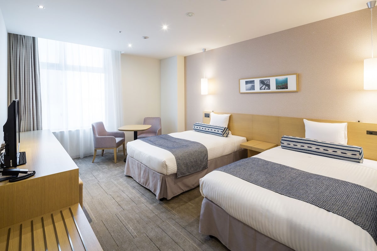 A hotel room with two beds, a desk, a chair, and a TV.