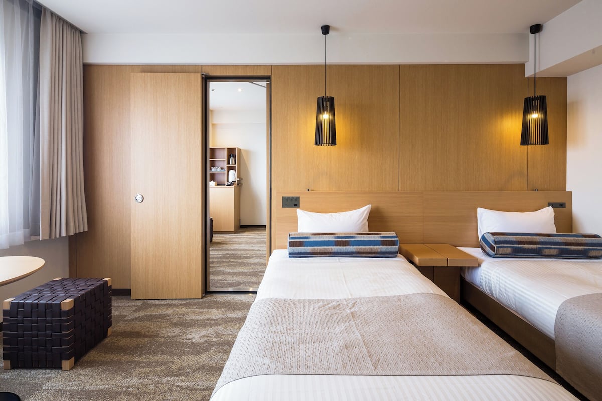 A hotel room with two beds, a wooden headboard, a small table, and a bench.