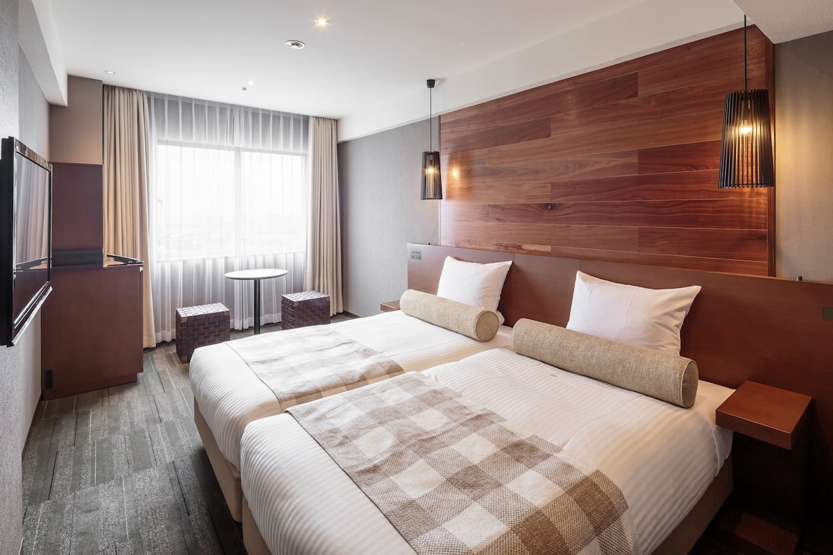 A hotel room with two beds, a wooden headboard, a television, a small table, and a window with curtains.