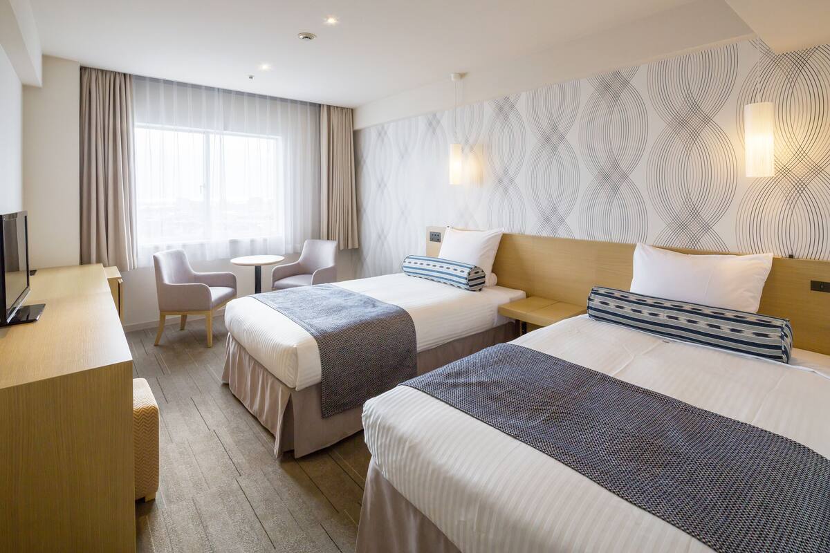 A hotel room with two beds, a desk, a chair, and a large window with curtains.