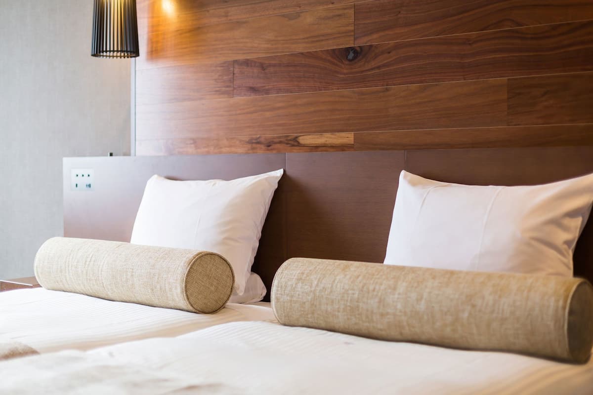A hotel room with a wooden headboard, two white pillows, and a rolled-up beige blanket on the bed.