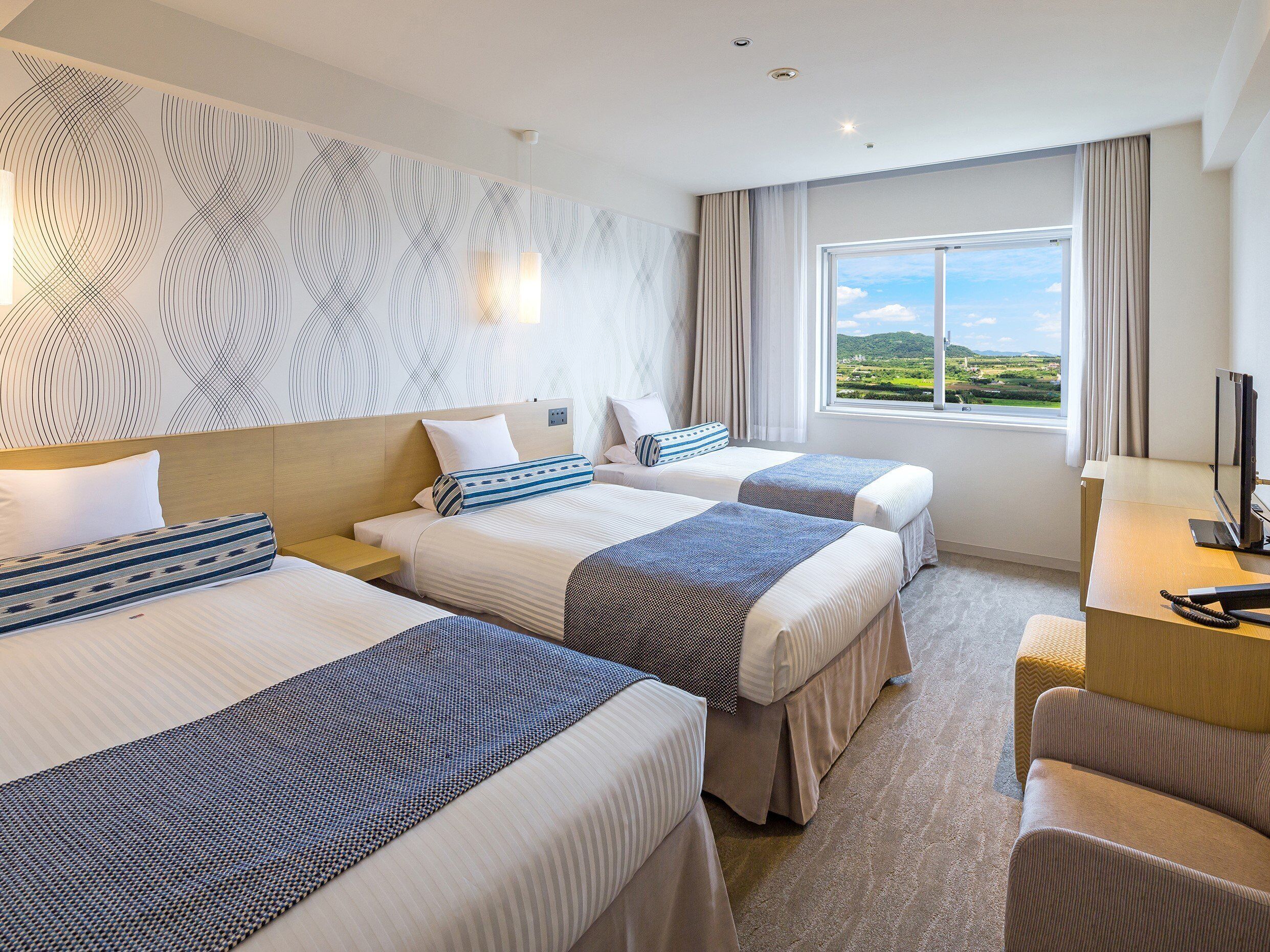 A hotel room with two beds, a desk, and a large window offering a view of the countryside.