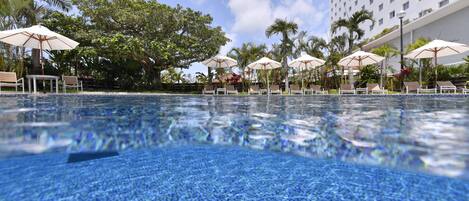 Piscina transparente com espreguiçadeiras e guarda-sóis, cercada por palmeiras e um prédio de hotel.