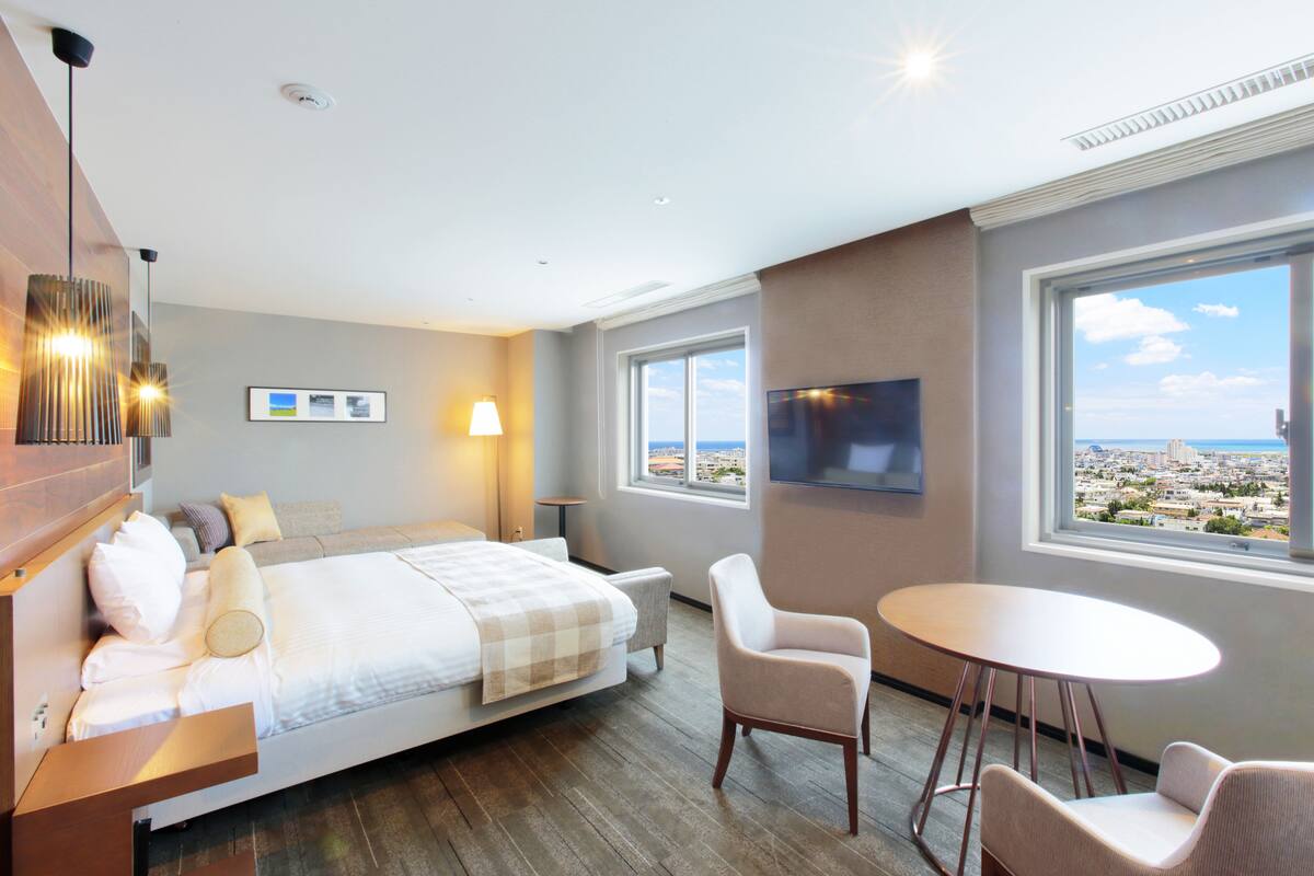 A hotel room with a bed, a desk, a chair, a table, a TV, and large windows with a city view.