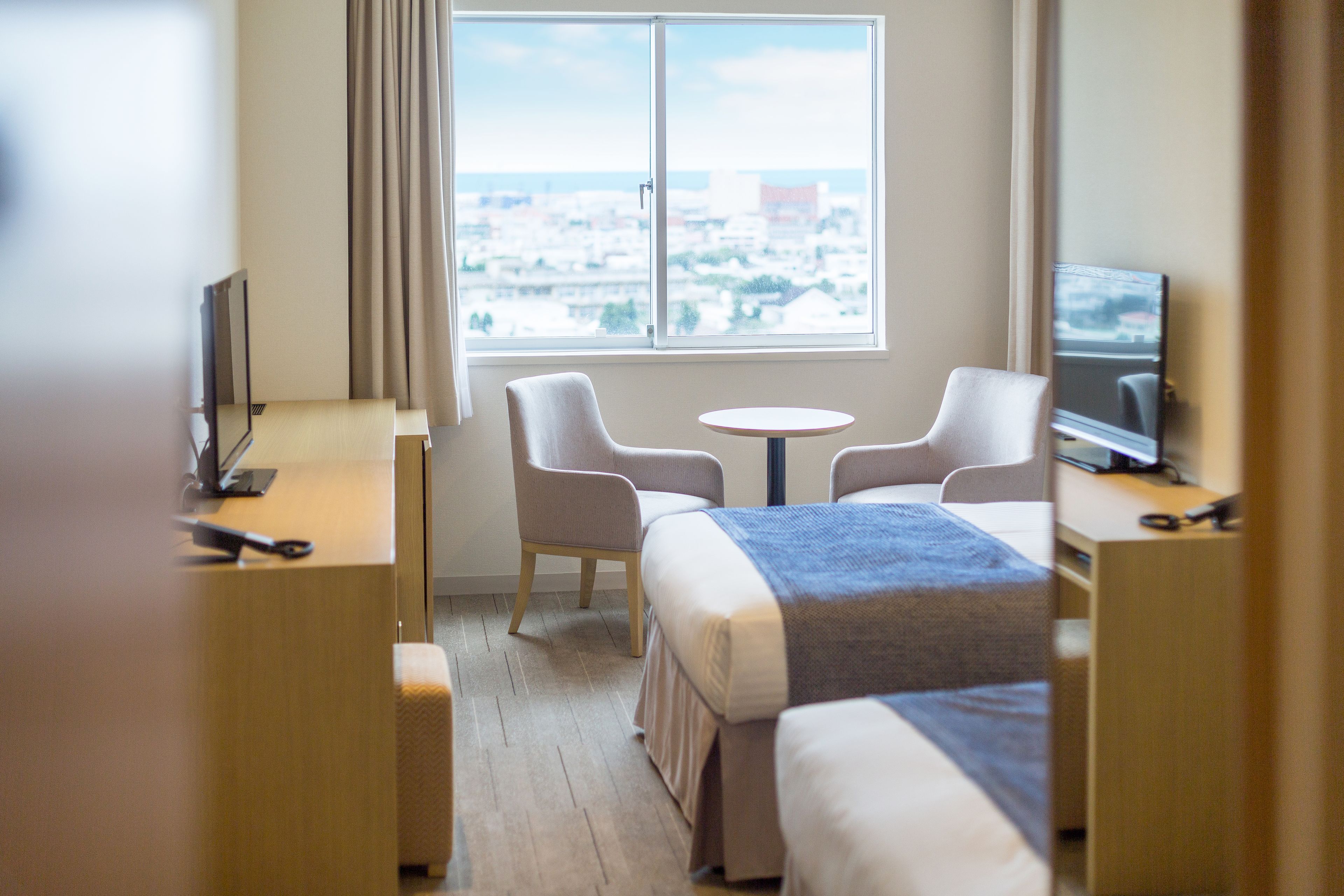A hotel room with two beds, a desk, a chair, a TV, and a window with a city view.