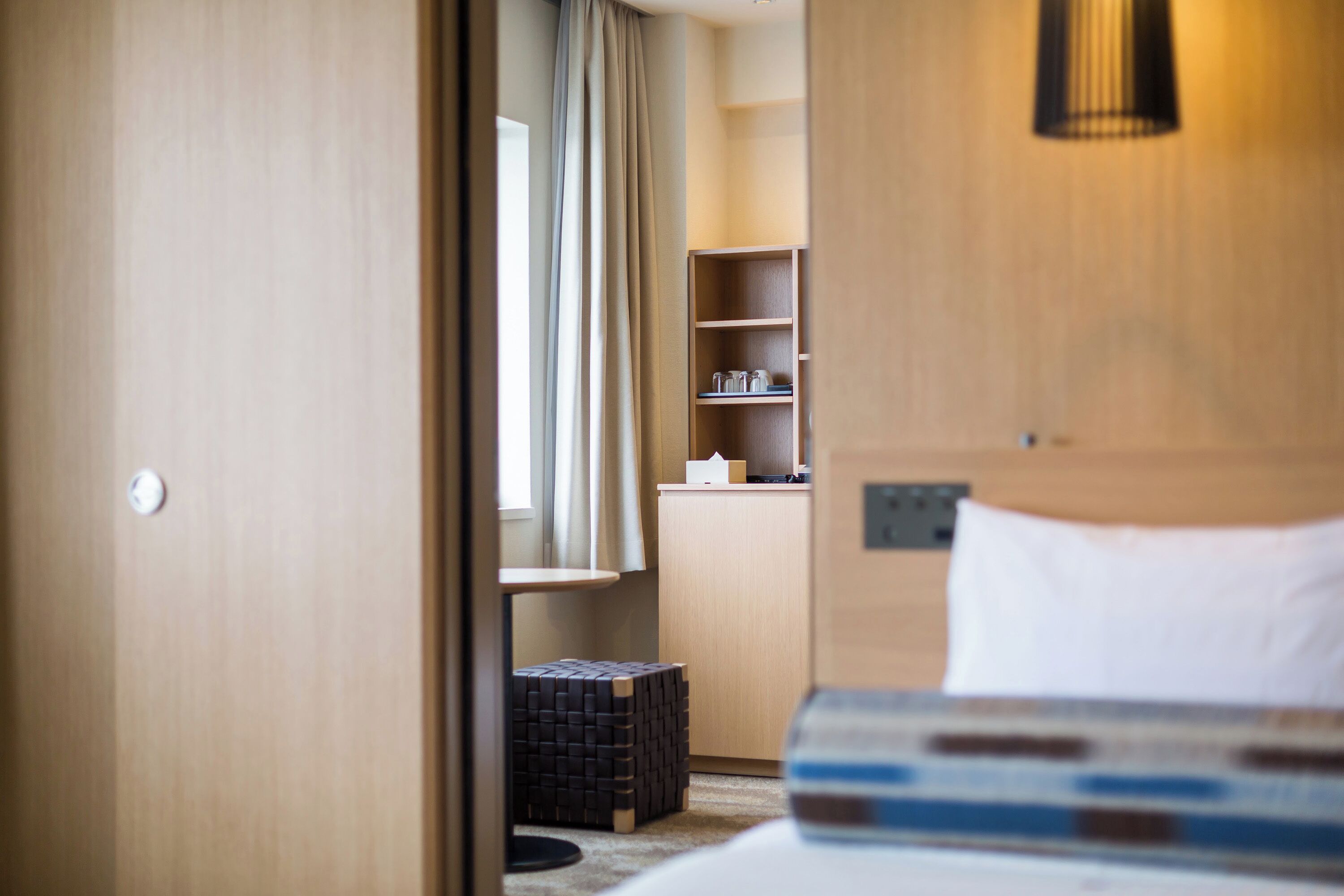 A hotel room with a bed, a small desk, a shelf with glasses, and a window with curtains.