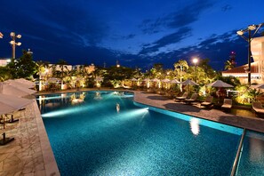 Zona de piscina nocturna con sillas de descanso y sombrillas, rodeada de palmeras y farolas.