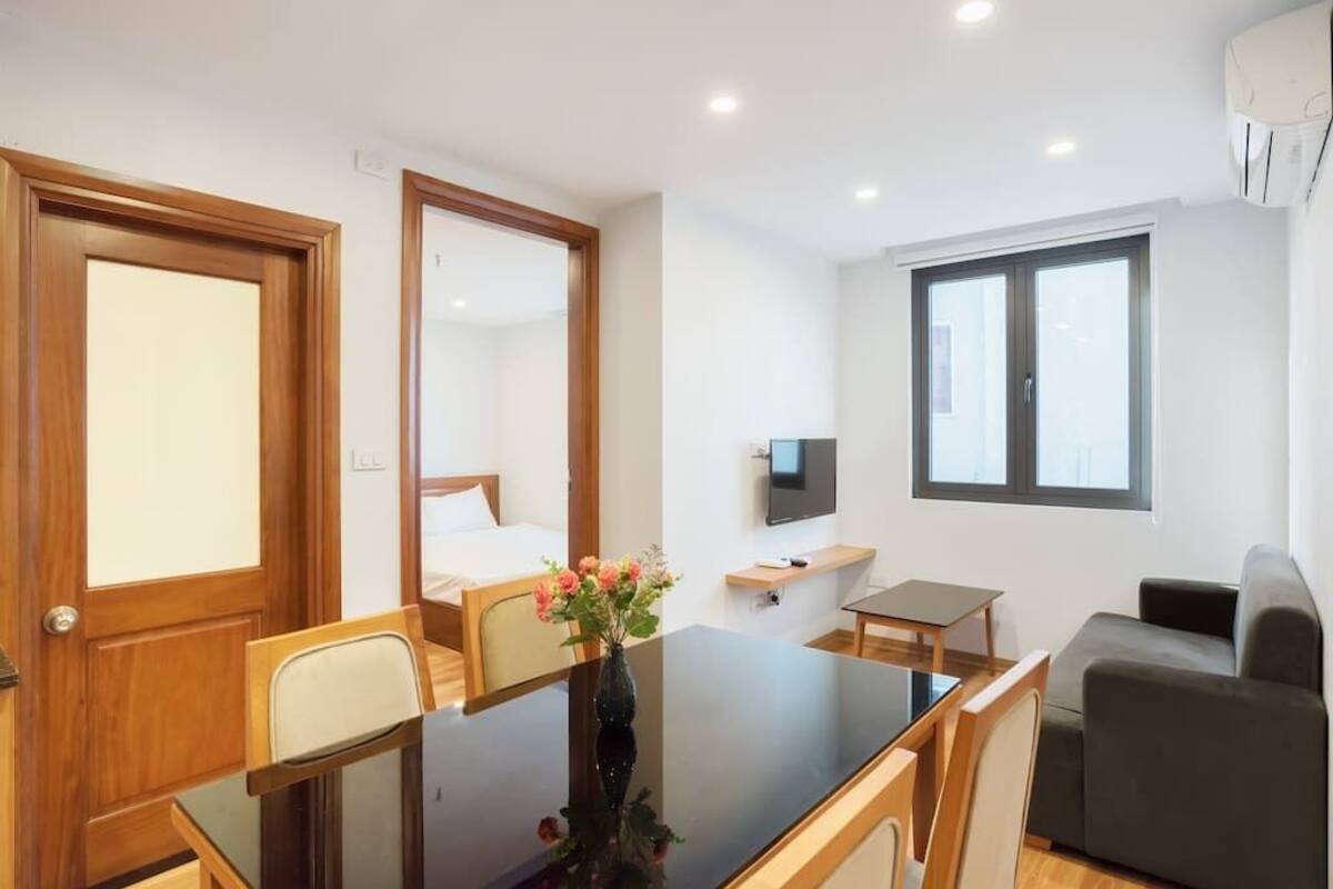 Una habitación de hotel moderna con una puerta de madera, una cama con ropa de cama blanca, una mesita con un jarrón de flores, un sofá gris y un televisor montado en la pared.