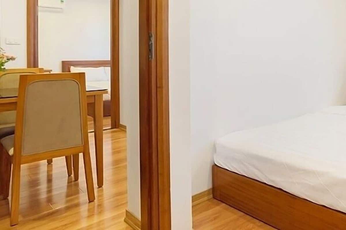 Una cama individual con cabecera de madera, un escritorio pequeño y una silla en una habitación con suelo de madera y paredes blancas.
