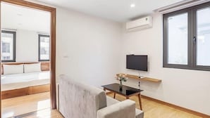 Una habitación moderna con una cama, un televisor, una mesita con un florero y ventanas amplias.