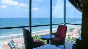 A room with a large window overlooking a beach and ocean. There are two red chairs and a small round table. - Le Hoang Beach Hotel (Da Nang)