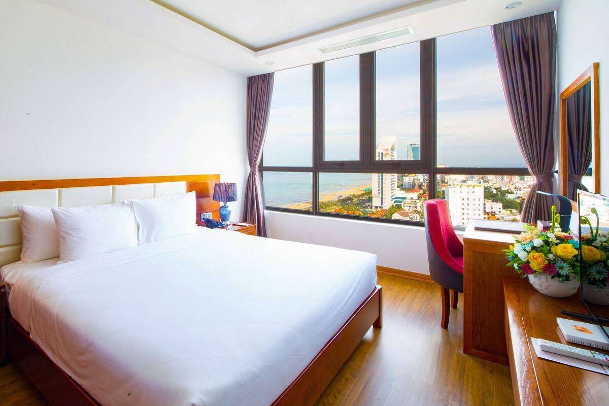 Hotelkamer met een groot bed, een bureau met stoel, uitzicht op stad en strand, en een raam met gordijnen.