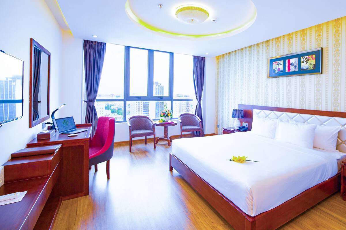 Een hotelkamer met een groot bed, een bureau met een laptop, twee stoelen, een kleine tafel met bloemen en een uitzicht op de stad door grote ramen.