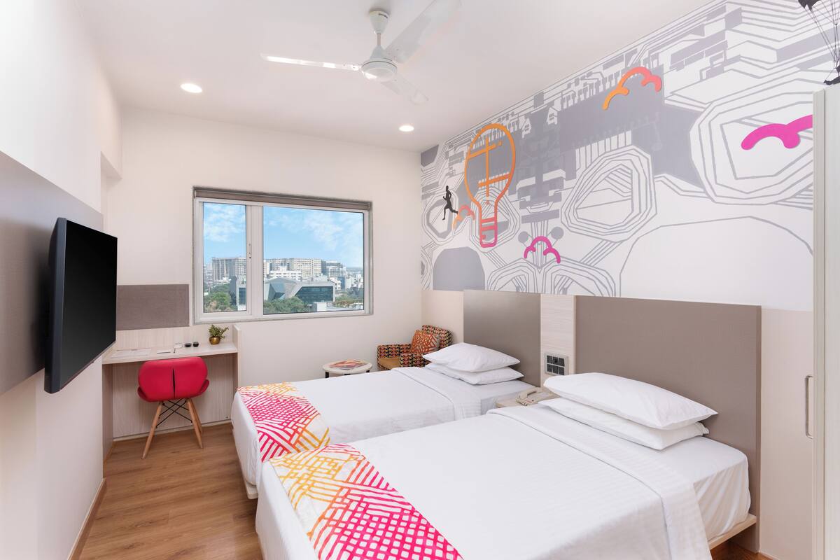 A modern hotel room with two beds, a desk, a TV, and a city view.