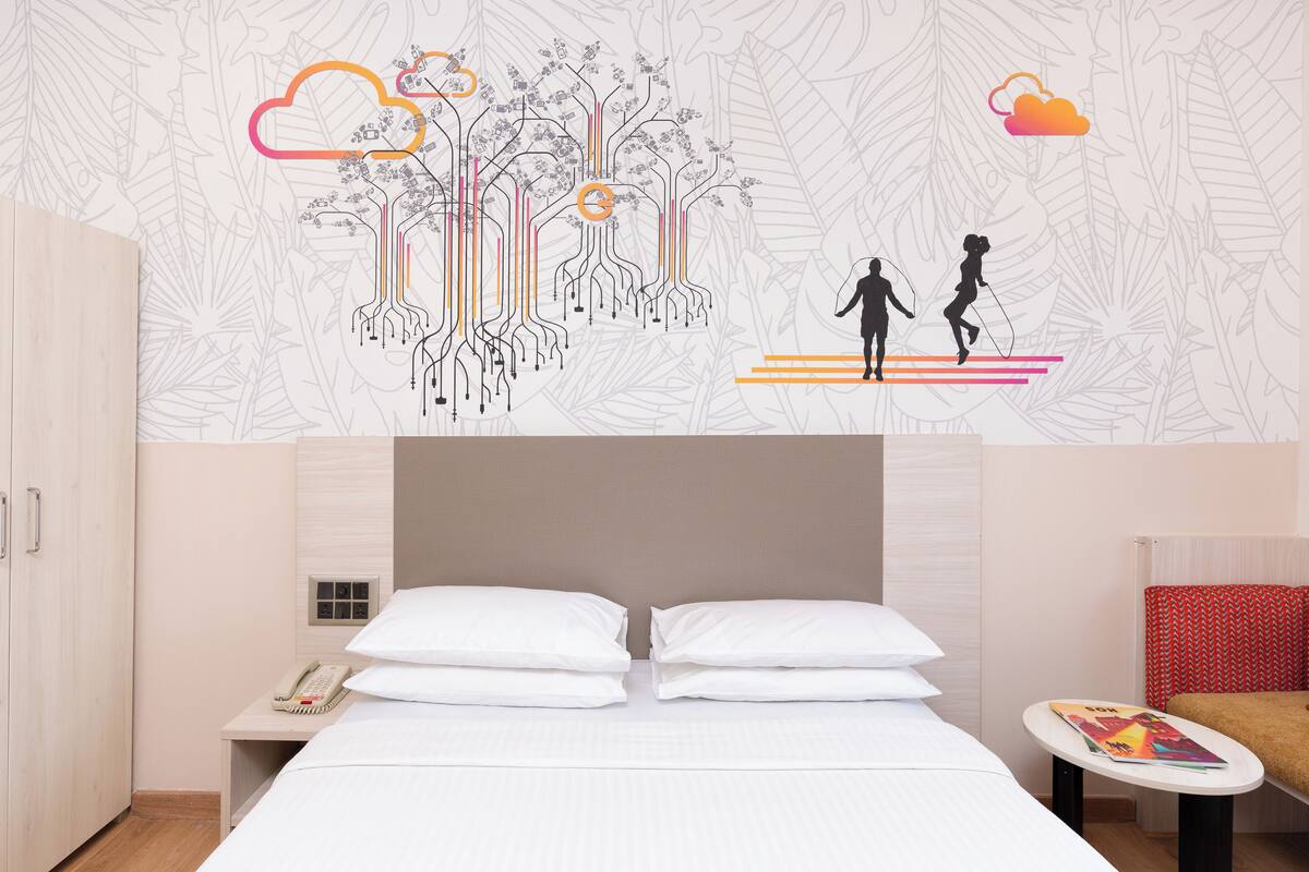 A hotel room with a bed, nightstands, a red chair, and a wall with a tree and cloud design.