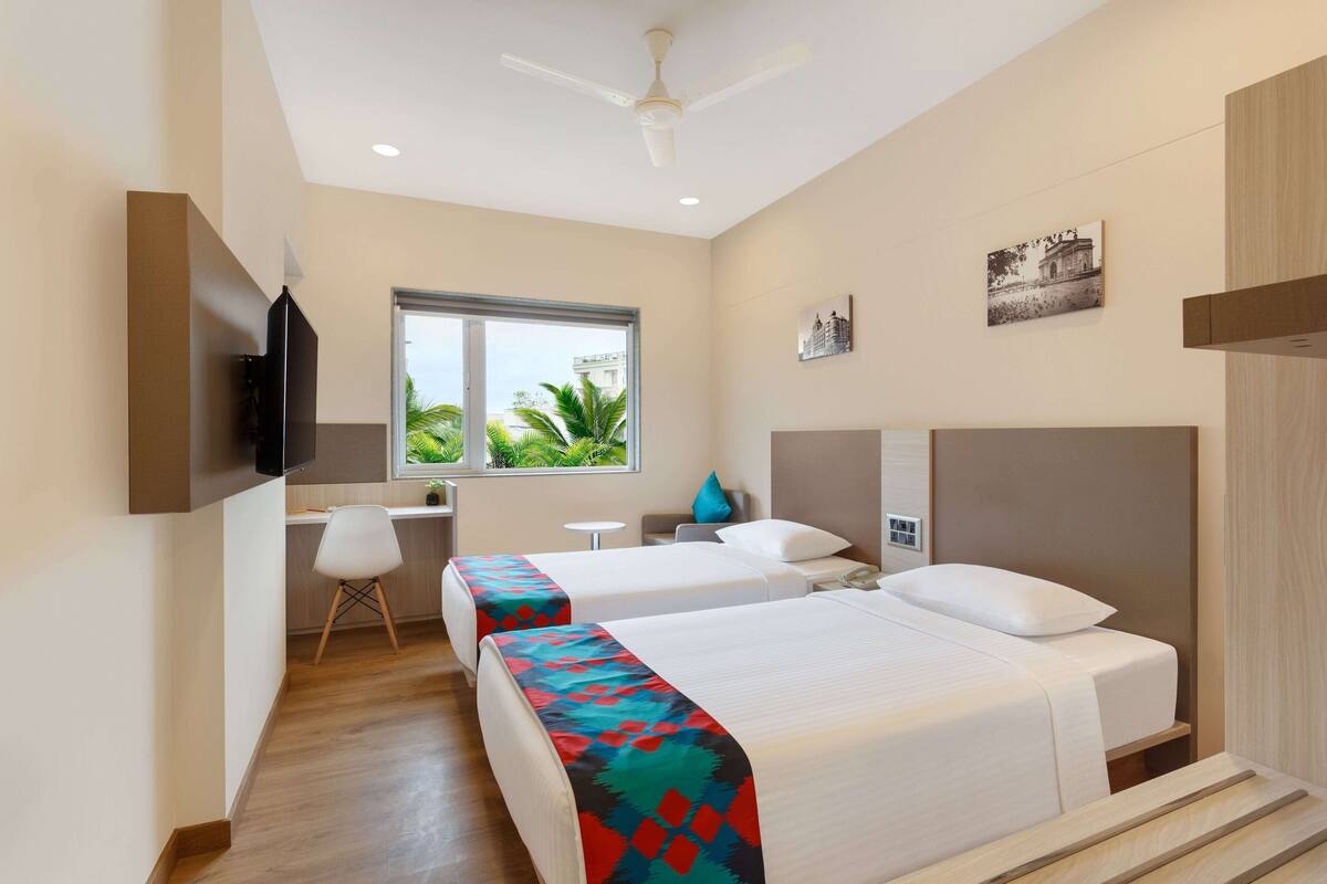 A hotel room with two beds, a desk, a chair, a TV, and a window with a view of greenery.
