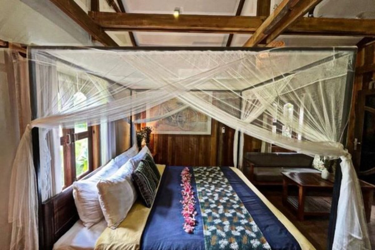 A room with a bed under a mosquito net, wooden beams, a window, and a table with a vase.