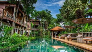 Un área de piscina con mobiliario de cubierta de madera, un pequeño pabellón cubierto y exuberante vegetación tropical.