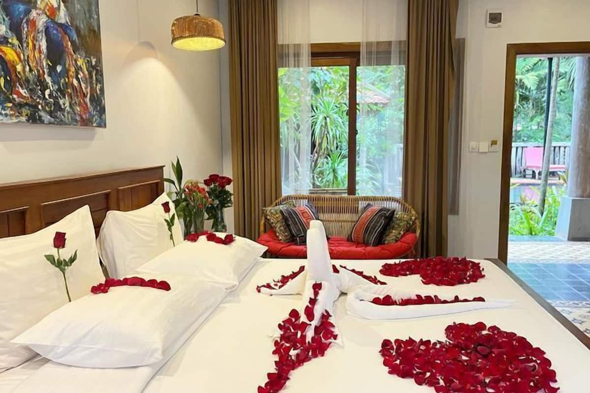 A hotel room with a bed adorned with rose petals, a painting on the wall, and a view of greenery through the window.