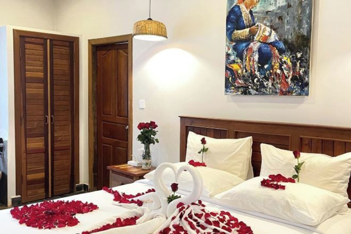 A hotel room with a bed adorned with rose petals and a heart-shaped arrangement, a painting on the wall, and wooden doors.