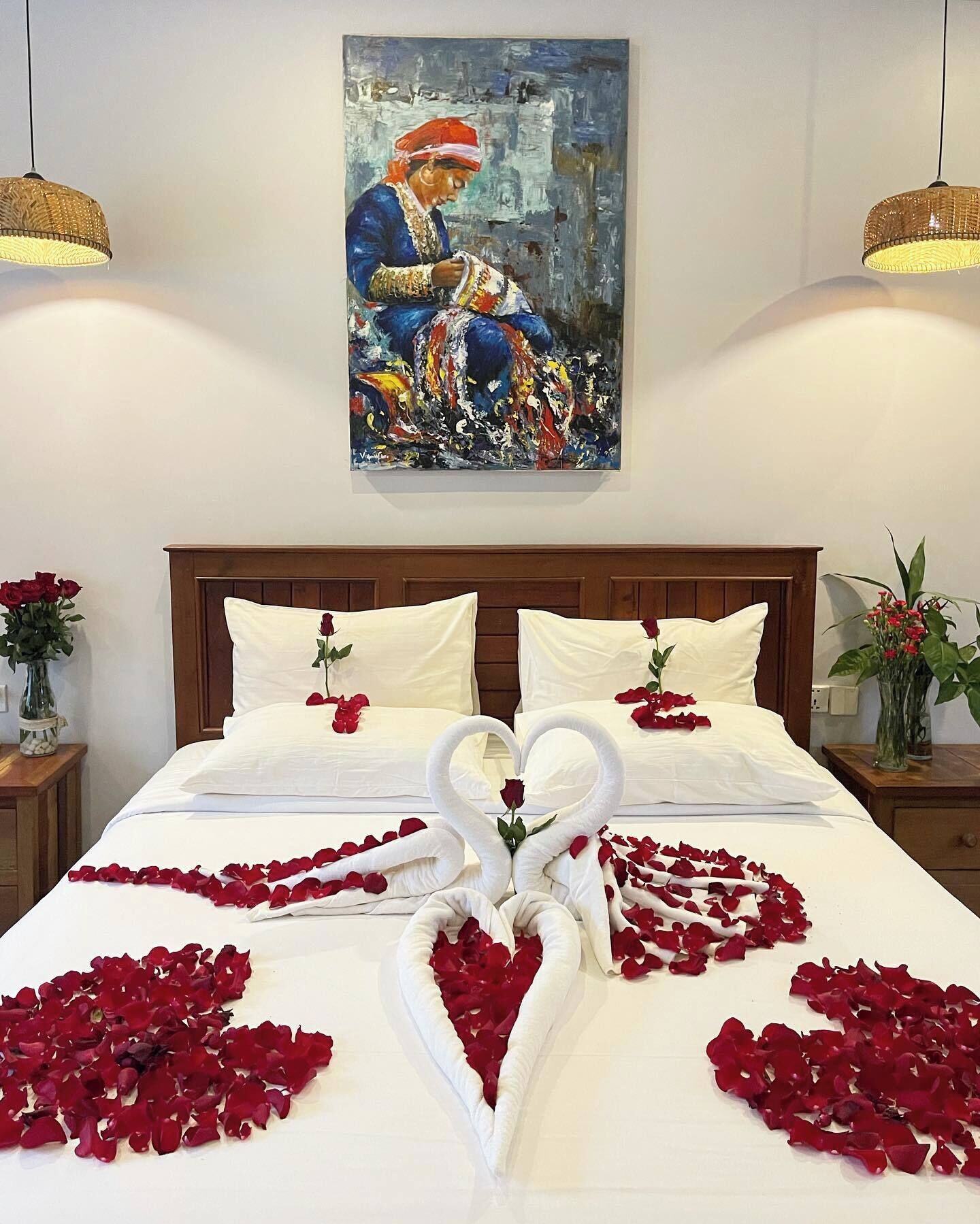 Una cama bien hecha con sábanas blancas, una toalla dispuesta en forma de corazón y pétalos de rosa esparcidos sobre la cama.
