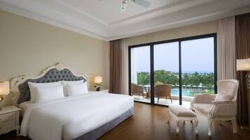 A spacious hotel room with a large bed, a sitting area with a chair and ottoman, a balcony with a view of the ocean, and a ceiling fan.