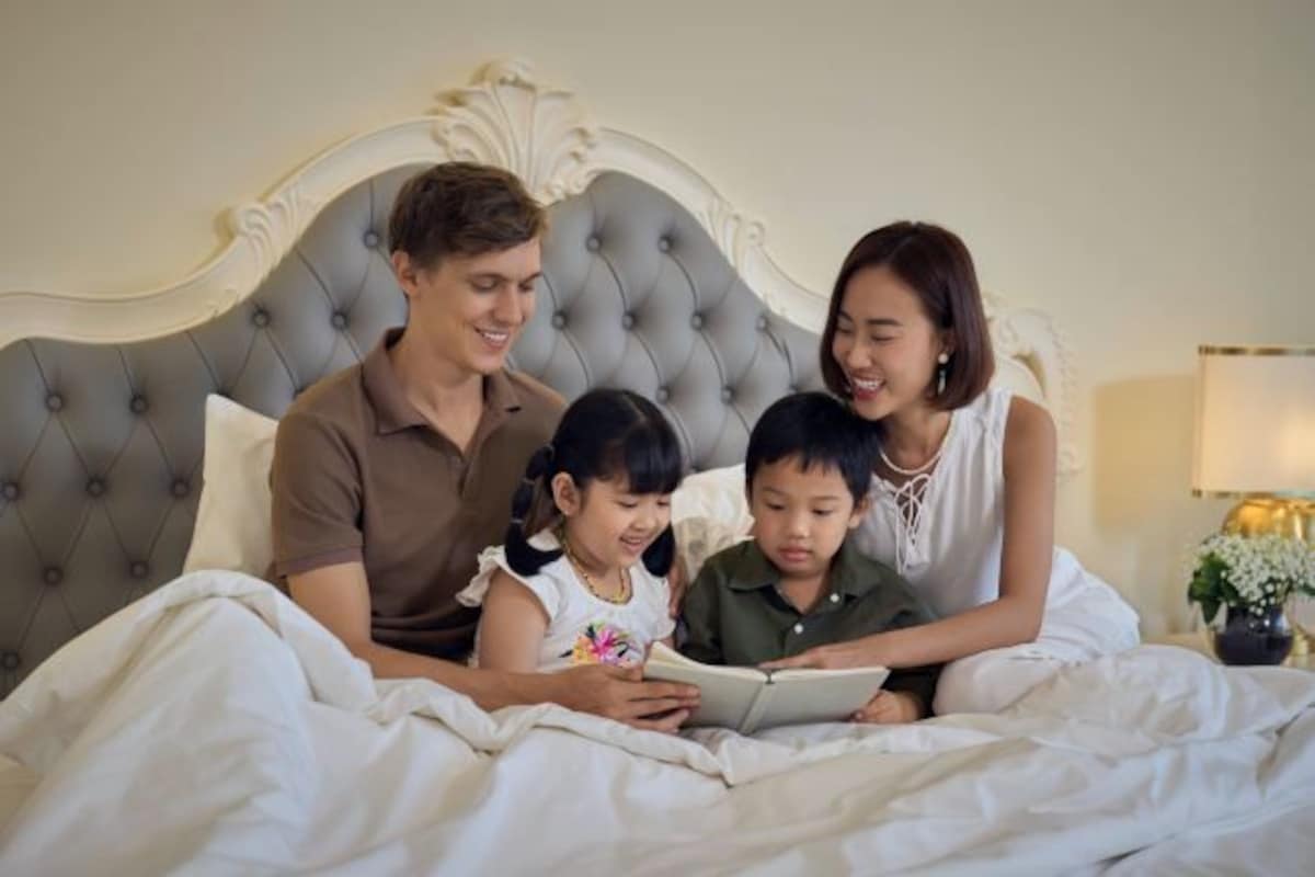 Une famille de quatre personnes est allongée sur un lit doté d’une tête de lit capitonnée, d’une lampe de chevet et agrémentée d’un arrangement floral.